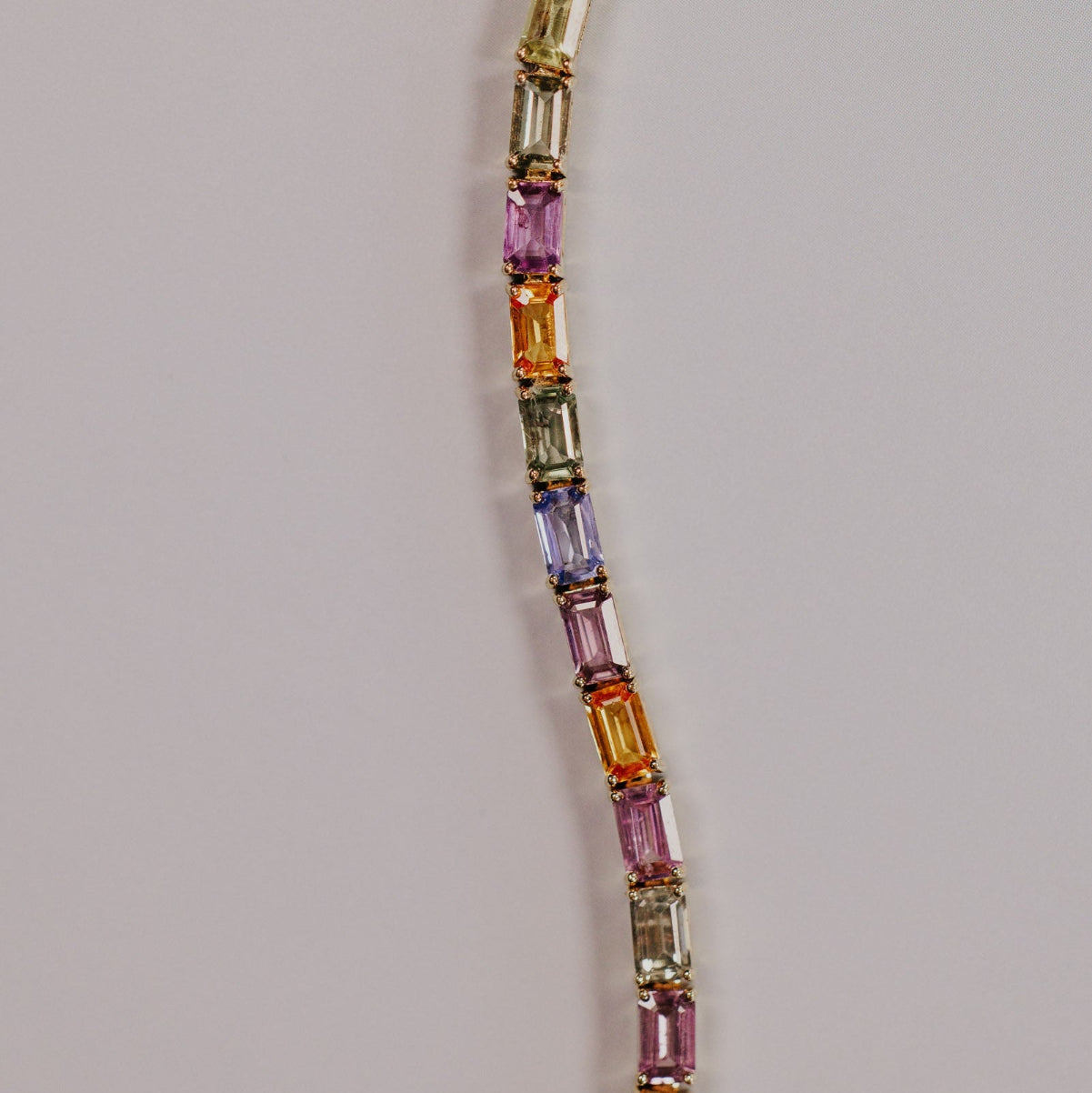 Close-up detail of the Pastel Emerald Cut Sapphire Tennis Bracelet in 14k yellow gold, highlighting the emerald cut natural sapphires in pastel pink, purple, blue, green, and orange tones in prong settings on a light gray surface.
