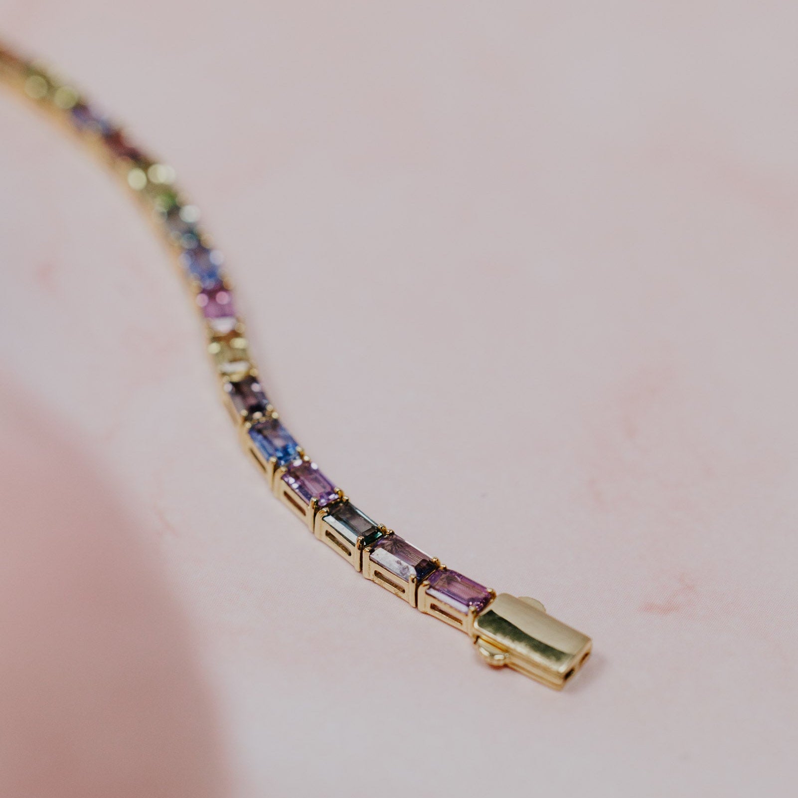 Close-up detail of the Pastel Emerald Cut Sapphire Tennis Bracelet in 14k yellow gold, highlighting the emerald cut pastel natural sapphires and gold clasp on a pink marble surface.
