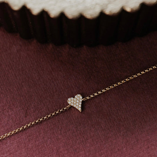 A delicate gold chain bracelet with a small pavé heart charm, displayed on a textured dark burgundy surface.
