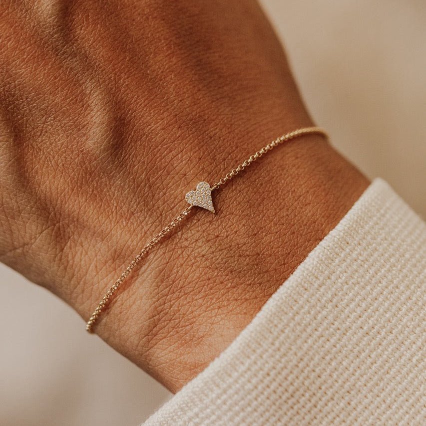 A delicate gold chain bracelet with a small, textured heart charm, worn on a person's wrist over a white knit cuff.