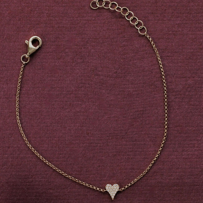 A delicate gold chain bracelet featuring a small pave-set heart charm, displayed on a textured dark burgundy fabric background.