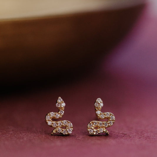 Gold snake-shaped stud earrings with pave diamonds on a dark background.