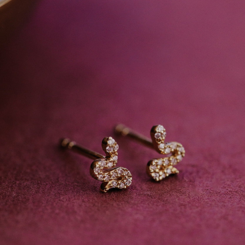 Gold snake shaped diamond stud earrings on a purple surface.
