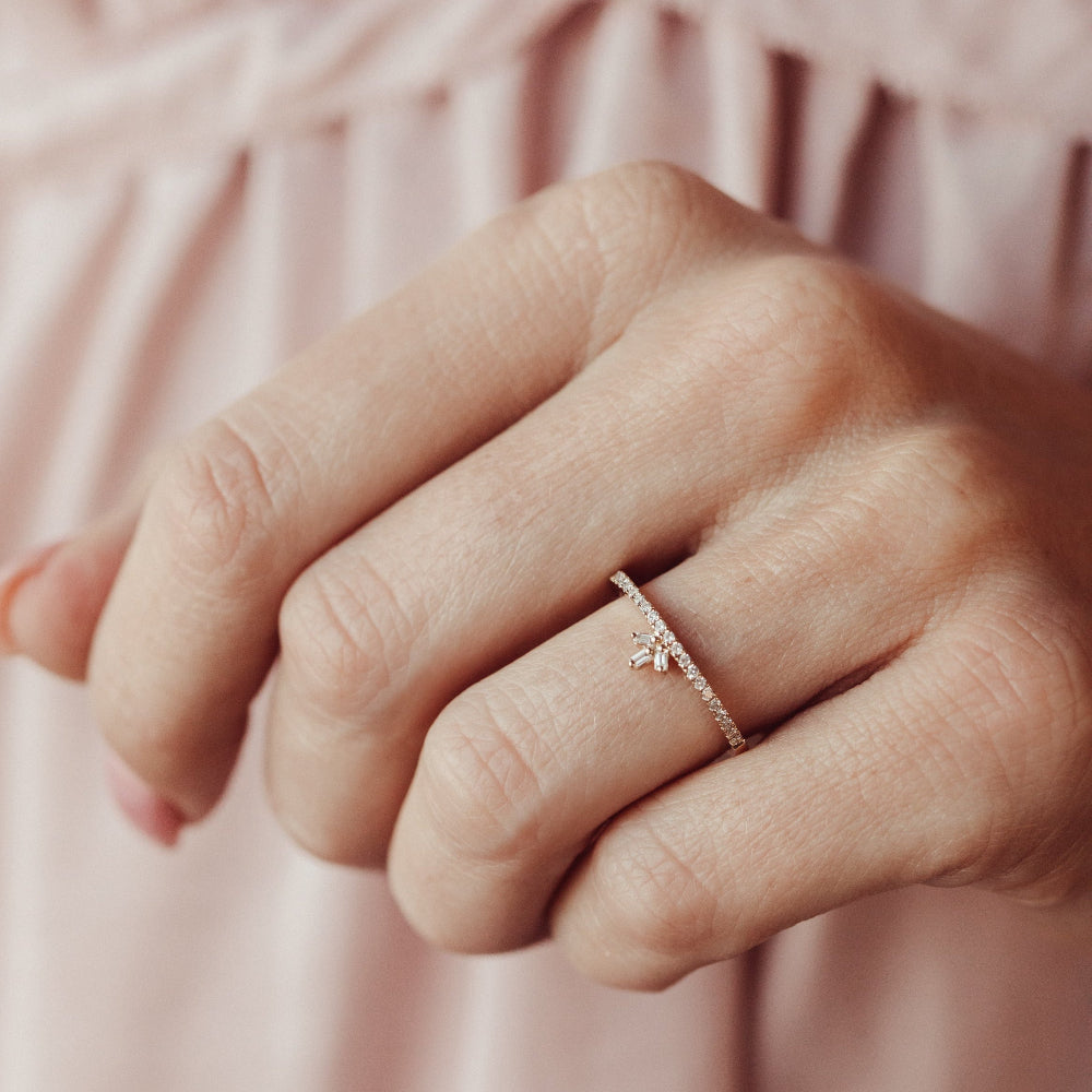 Close-up of a hand wearing a delicate diamond wedding band on a soft pink background.