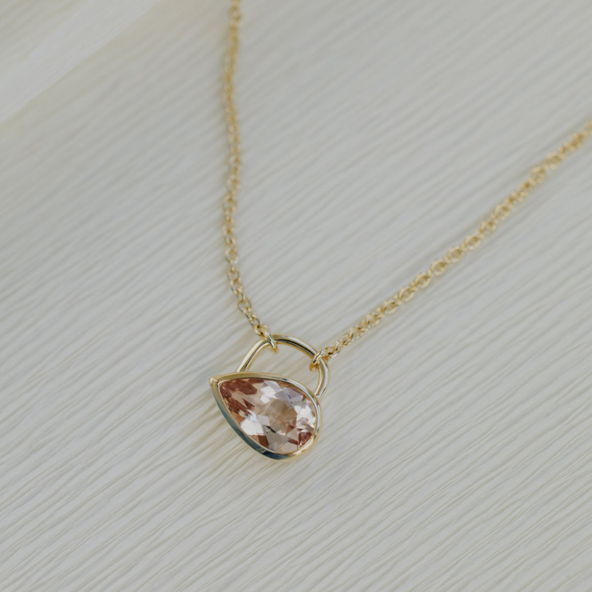 A light pink Pear shaped morganite stone in a bezel setting on a textured white background.