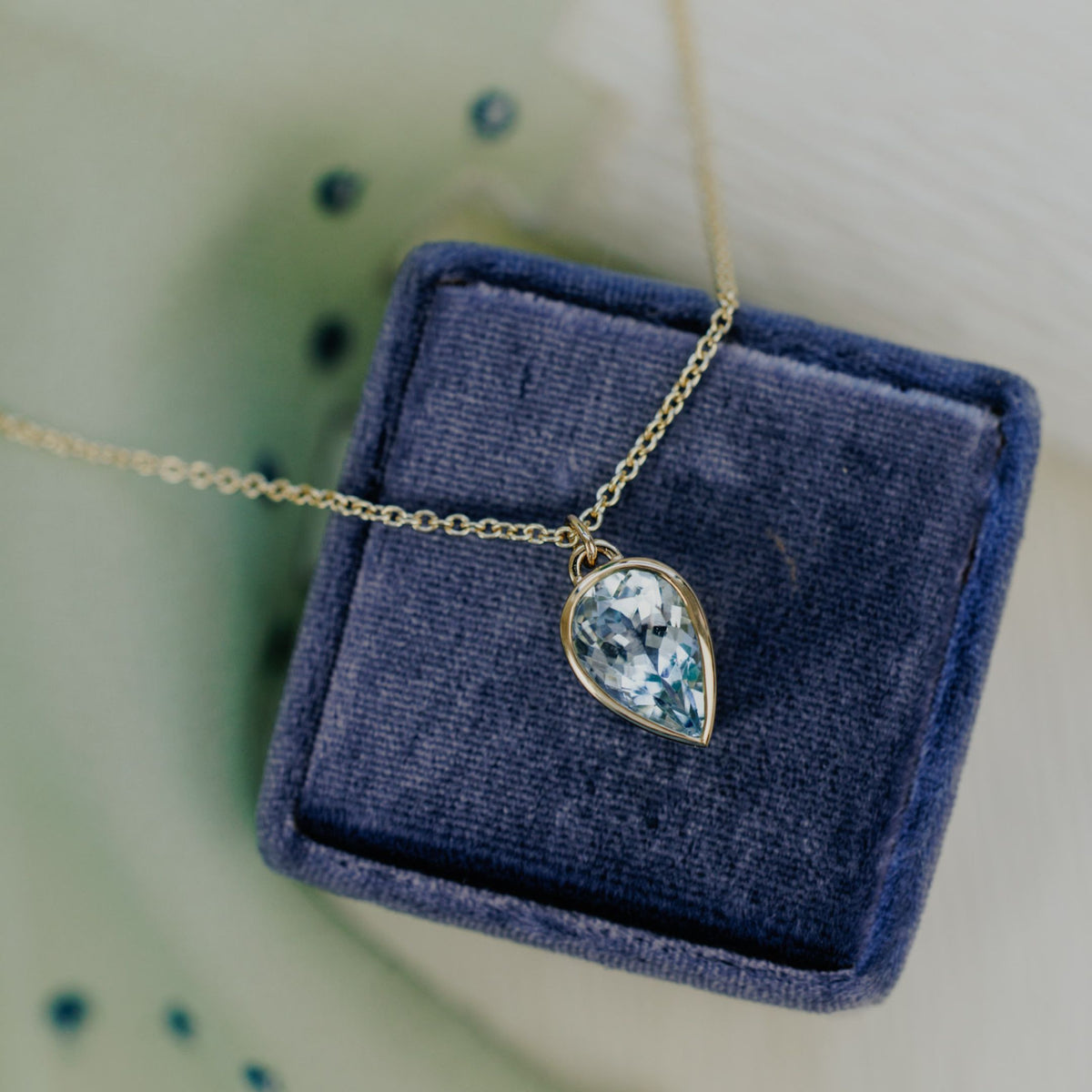 A 14k yellow gold pear-shaped aquamarine necklace in a bezel set design on a blue jewelry box with a textured background.