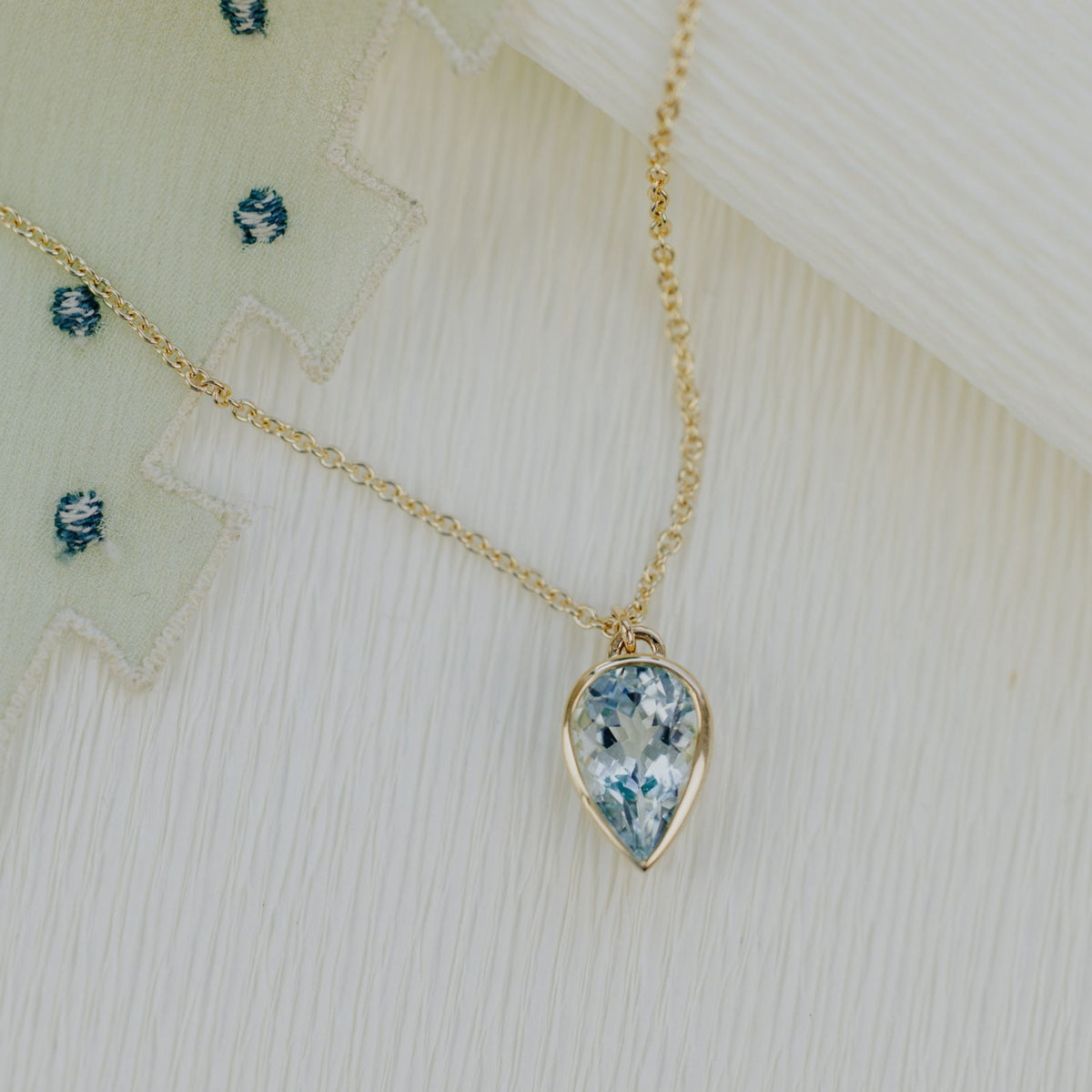 A 14k yellow gold pear-shaped aquamarine necklace in a bezel set design on a textured background.