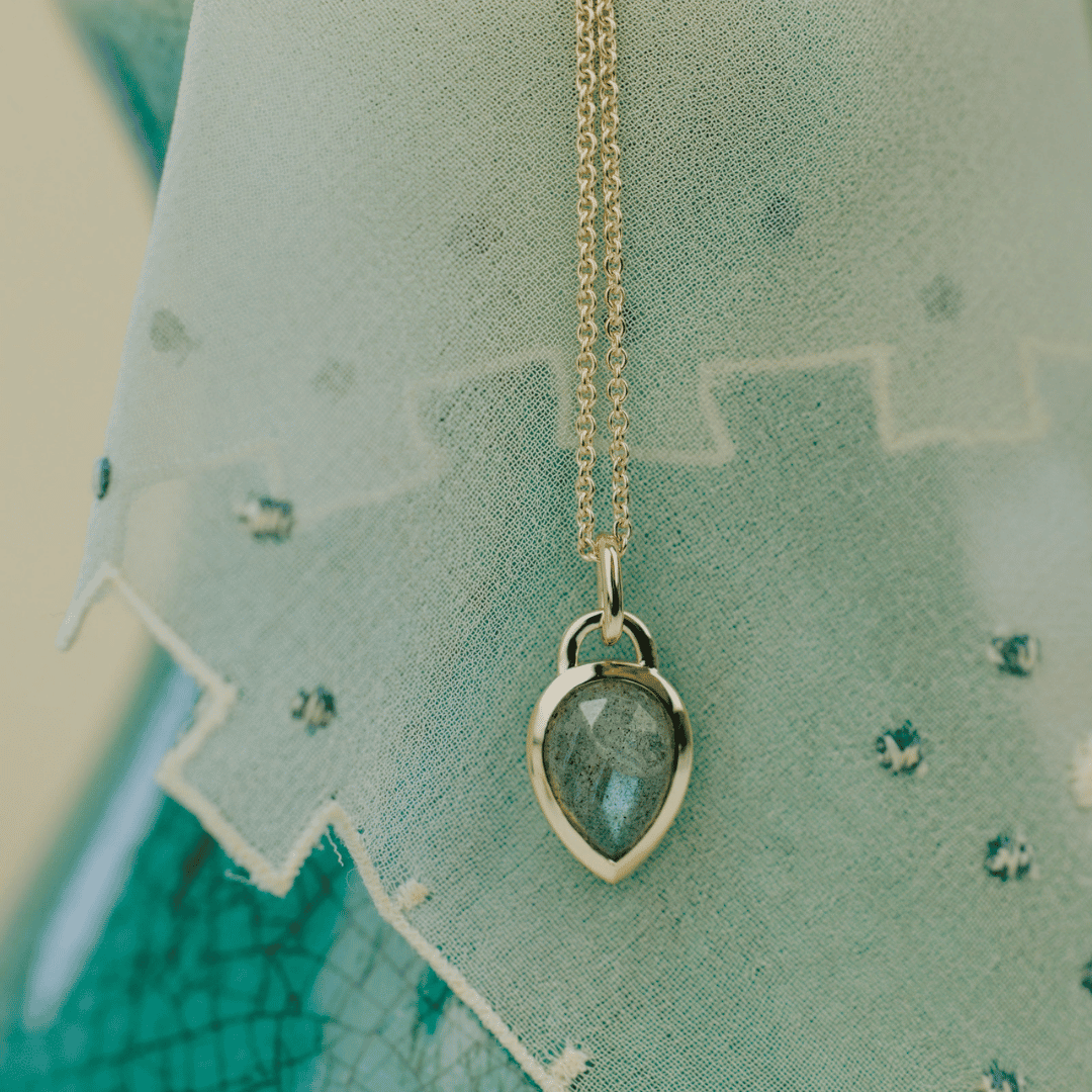 A 14k yellow gold pear shaped labradorite necklace in a bezel setting featured on a textured background. 