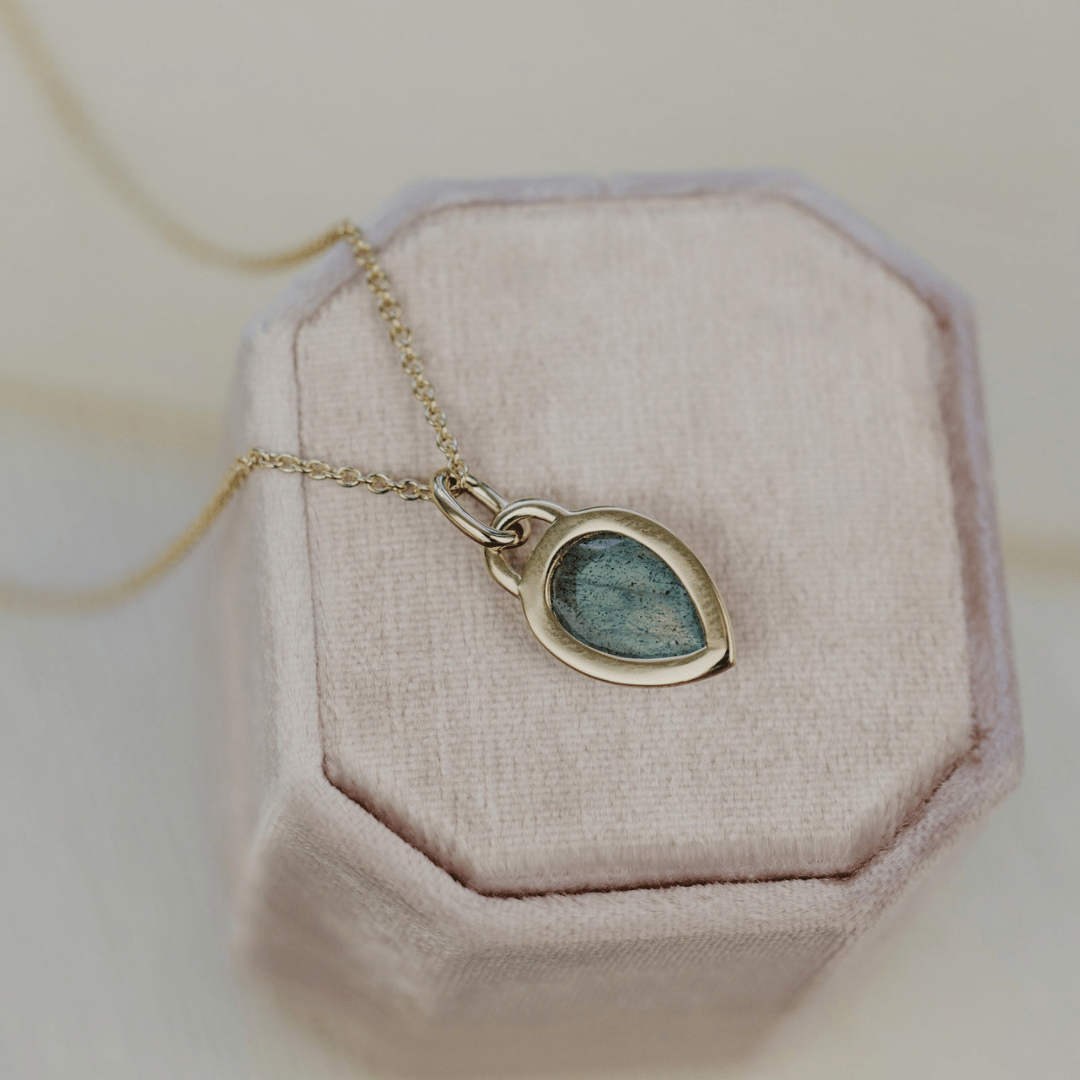 The back side of a 14k yellow gold pear shaped labradorite necklace in a bezel setting featured on a light pink jewelry box. 
