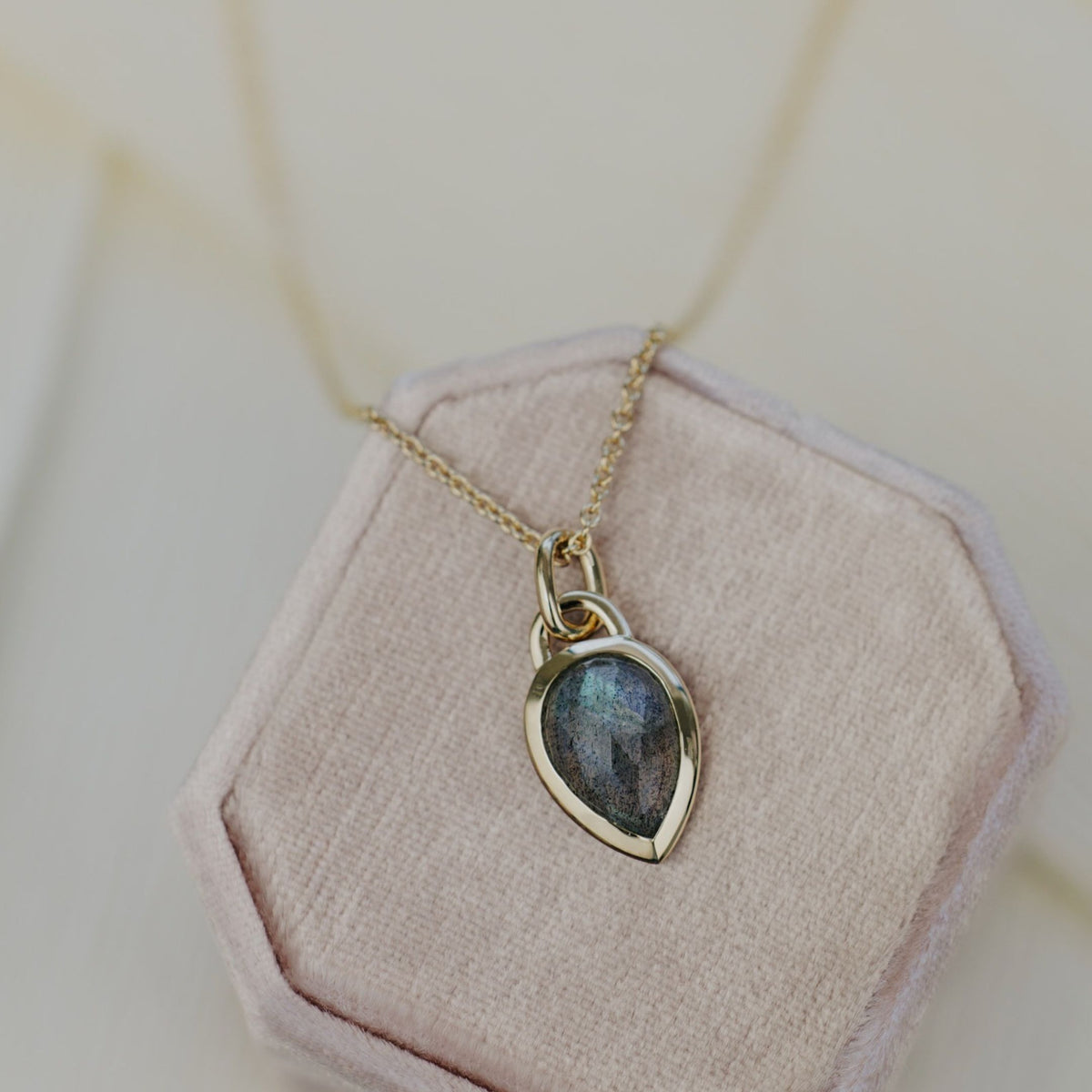 A 14k yellow gold pear shaped labradorite necklace in a bezel setting featured on a light pink jewelry box. 
