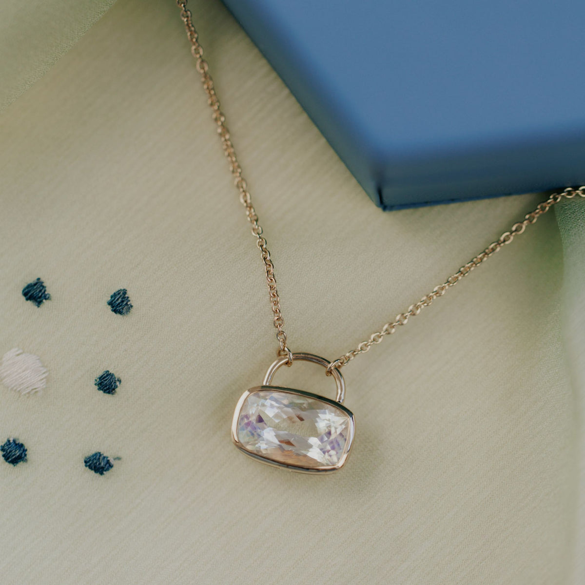 A 14k yellow gold cushion cut moonstone necklace in a bezel setting with a lock style design on a textured background.
