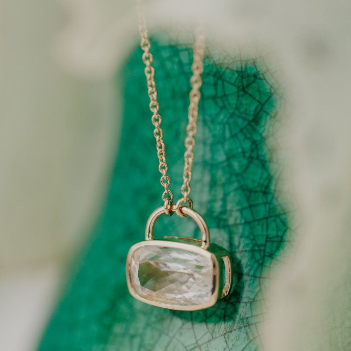 A 14k yellow gold cushion cut moonstone necklace in a bezel setting with a lock style design on a textured background.