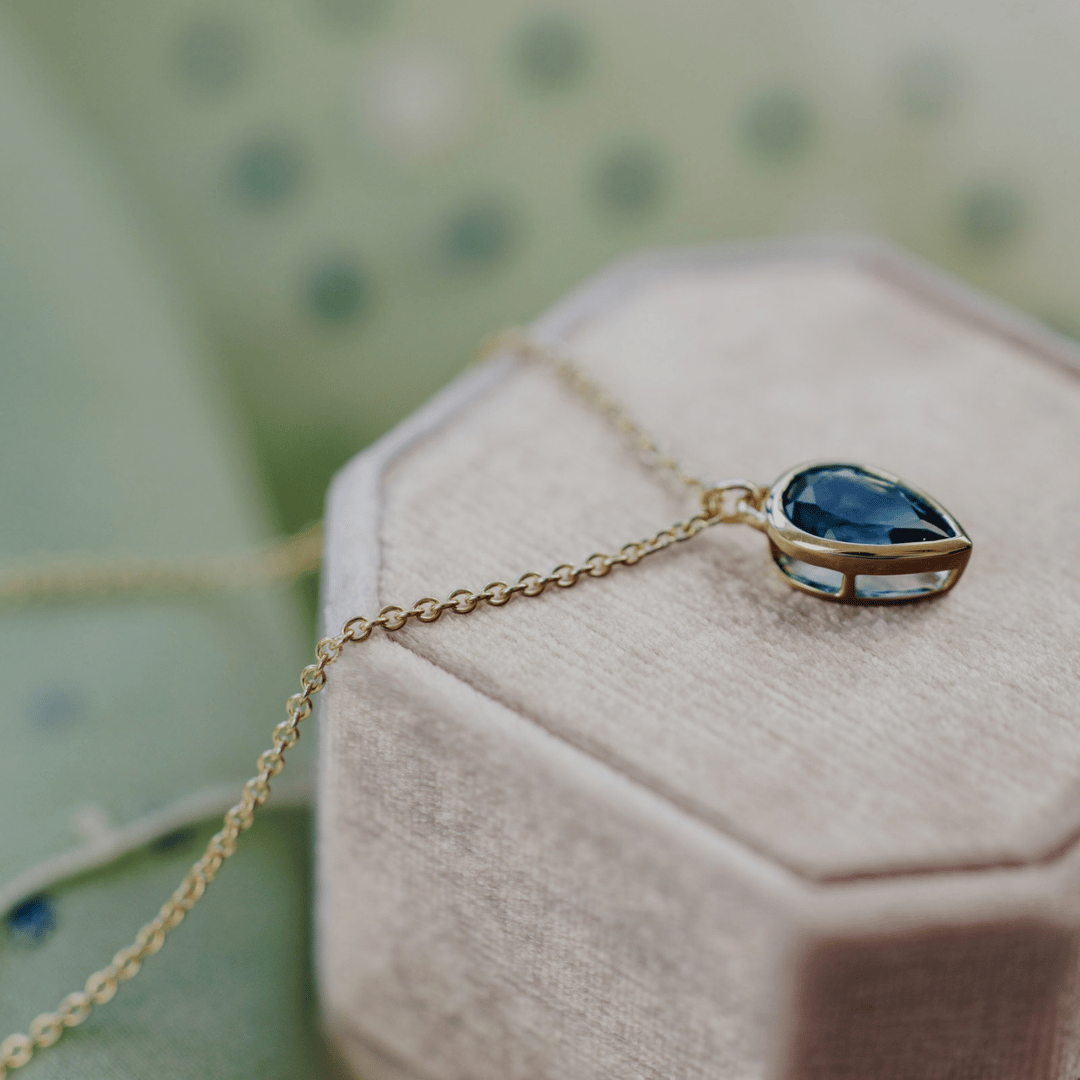 A 14k gold necklace with a pear shaped blue topaz stone in a bezel set design on a pink jewelry box with a soft background. 