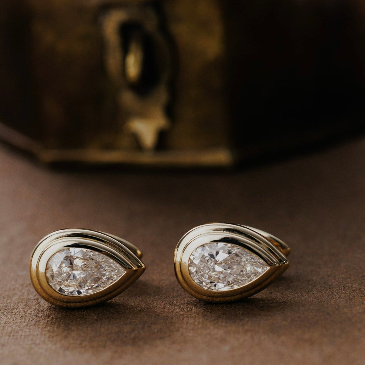 Gold earrings with pear-shaped lab grown diamonds on a brown surface with a vintage box in the background.