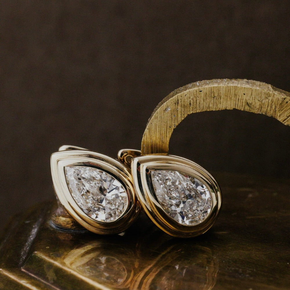 Gold earrings with pear-shaped lab grown diamonds on a reflective surface.