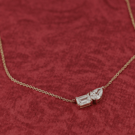 A 14k gold necklace featuring two lab grown diamonds in pear and emerald shapes, displayed on a textured red surface.