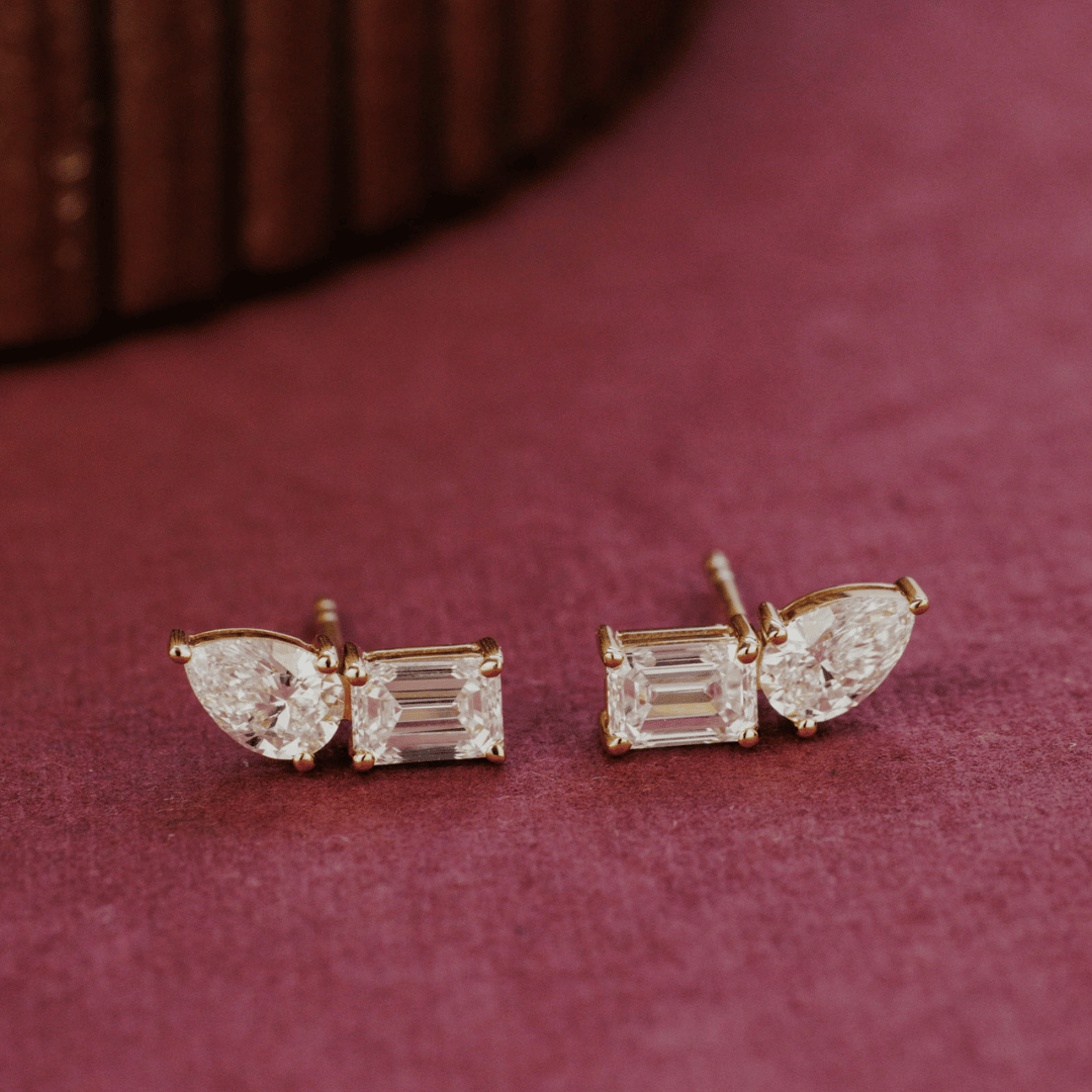 14k gold diamond earrings emerald and pear shaped set in prongs showcased on a textured red surface. 
