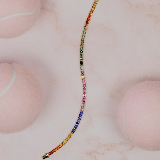 The Rainbow Emerald Cut Sapphire Tennis Bracelet in 14k yellow gold, featuring 13.68 cttw emerald cut natural sapphires in a spectrum of vibrant colors secured in prong settings, shown unclasped on a pink marble surface.
