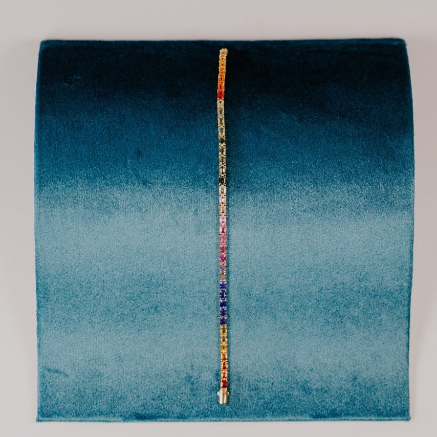 The Rainbow Emerald Cut Sapphire Tennis Bracelet in 14k yellow gold, featuring 13.68 cttw emerald cut natural sapphires in a spectrum of vibrant colors secured in prong settings, displayed upright on a teal velvet bracelet stand.
