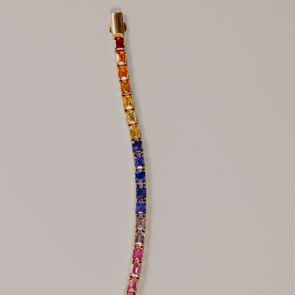 The Rainbow Emerald Cut Sapphire Tennis Bracelet in 14k yellow gold, featuring 13.68 cttw emerald cut natural sapphires in a spectrum of vibrant colors secured in prong settings, shown unclasped on a light gray surface with the clasp visible.
