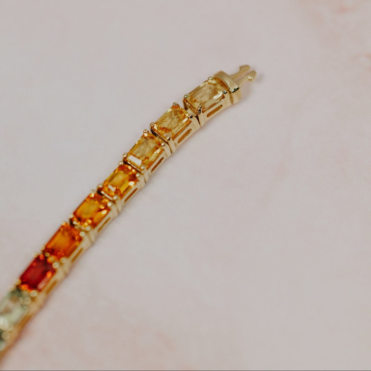 Close-up detail of the Rainbow Emerald Cut Sapphire Tennis Bracelet in 14k yellow gold, highlighting the yellow, orange, and red natural sapphires near the clasp end on a pink marble surface.