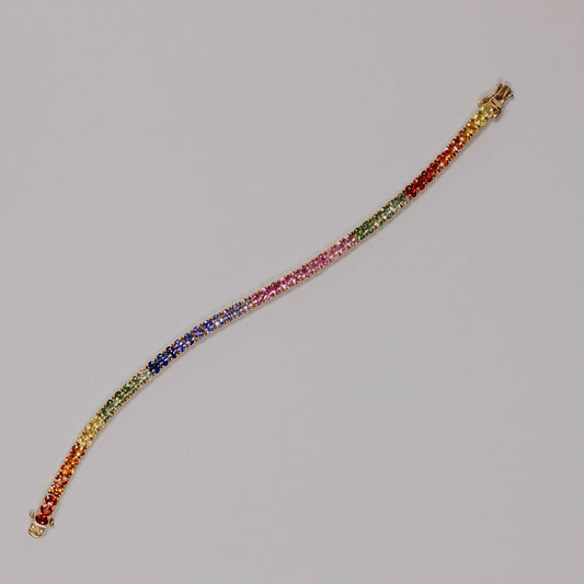 The Rainbow Oval Sapphire Tennis Bracelet in 14k yellow gold, featuring 11.25 cttw oval natural sapphires in a spectrum of vibrant colors secured in prong settings, shown unclasped on a light gray surface.
