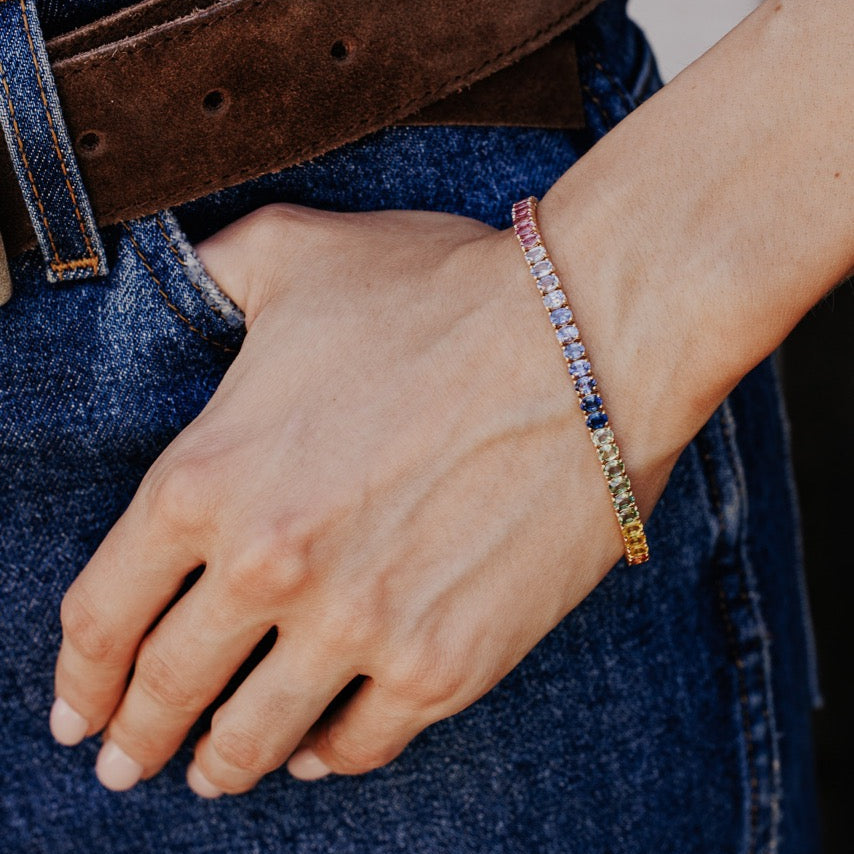 The Rainbow Oval Sapphire Tennis Bracelet in 14k yellow gold worn on the wrist against a casual denim outfit.
