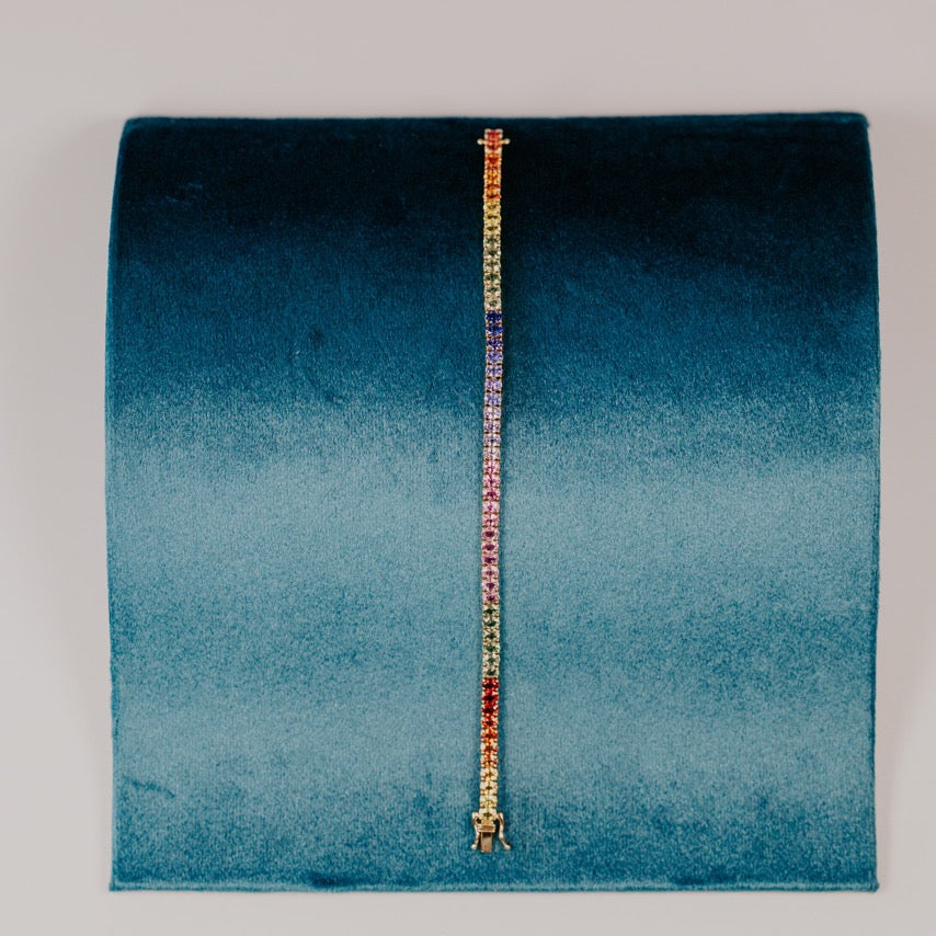The Rainbow Oval Sapphire Tennis Bracelet in 14k yellow gold, featuring 11.25 cttw oval natural sapphires in a spectrum of vibrant colors secured in prong settings, displayed upright on a teal velvet bracelet stand.
