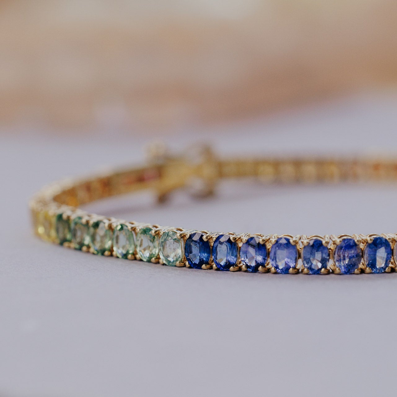 Close-up detail of the Rainbow Oval Sapphire Tennis Bracelet in 14k yellow gold, highlighting the graduated blue, teal, and green oval natural sapphires in prong settings on a light surface.

