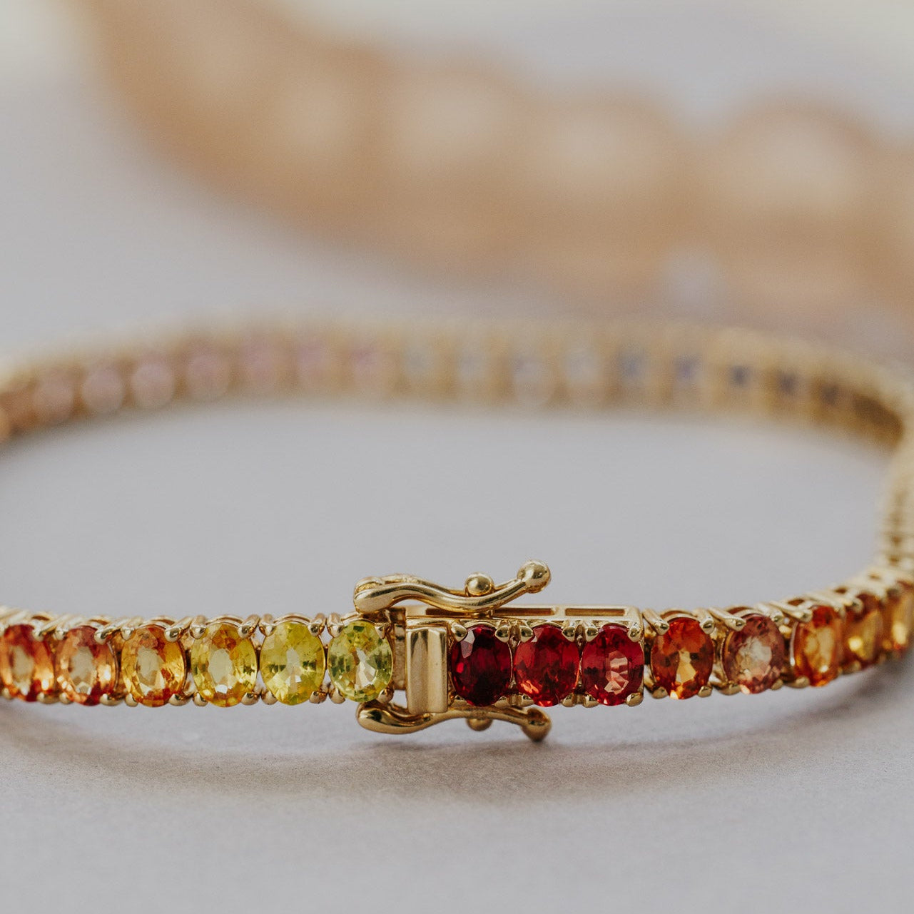 Close-up detail of the Rainbow Oval Sapphire Tennis Bracelet in 14k yellow gold, highlighting the red, orange, and yellow oval natural sapphires near the clasp on a light surface.
