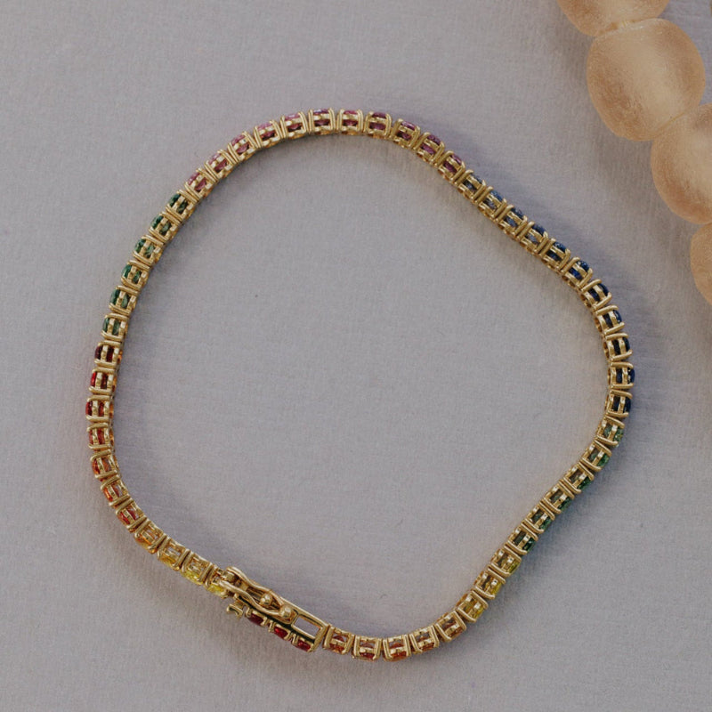 The Rainbow Oval Sapphire Tennis Bracelet in 14k yellow gold shown from the reverse side, displaying the full prong setting structure and the complete spectrum of multicolored oval natural sapphires on a light gray surface.