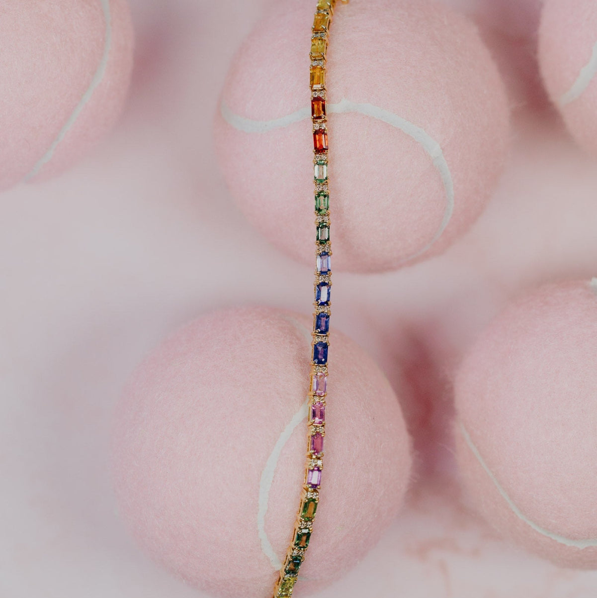 The Rainbow Sapphire with Diamond Accent Tennis Bracelet in 14k yellow gold, featuring 8.77 cttw emerald cut natural sapphires in a spectrum of vibrant colors with natural diamond accents secured in prong settings, shown unclasped across pink tennis balls.
