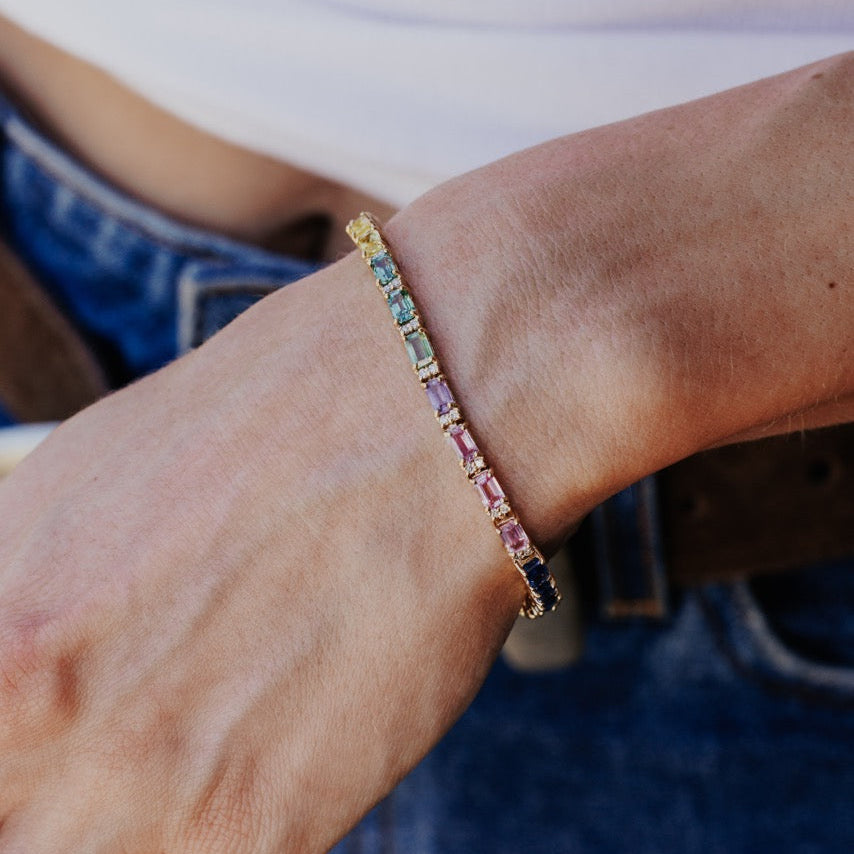The Rainbow Sapphire with Diamond Accent Tennis Bracelet in 14k yellow gold worn on the wrist, showing the emerald cut natural sapphires and diamond accents against a casual denim and white top outfit.
