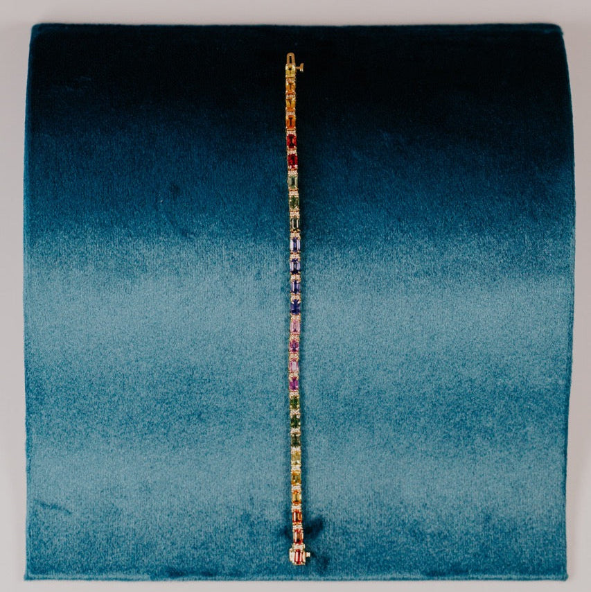 The Rainbow Sapphire with Diamond Accent Tennis Bracelet in 14k yellow gold, featuring 8.77 cttw emerald cut natural sapphires in a spectrum of vibrant colors with natural diamond accents secured in prong settings, displayed upright on a teal velvet bracelet stand.
