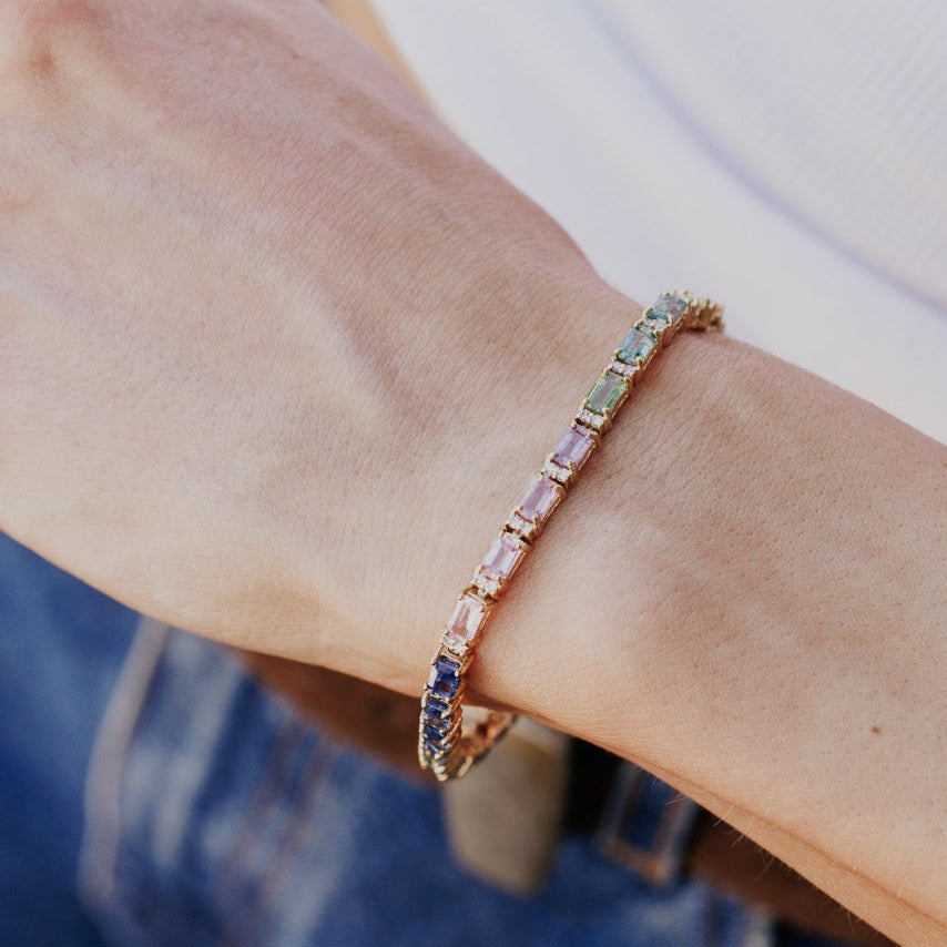 The Rainbow Sapphire with Diamond Accent Tennis Bracelet in 14k yellow gold worn on the wrist, showing the reverse side of the emerald cut natural sapphires and diamond accents against a casual denim and white top outfit.
