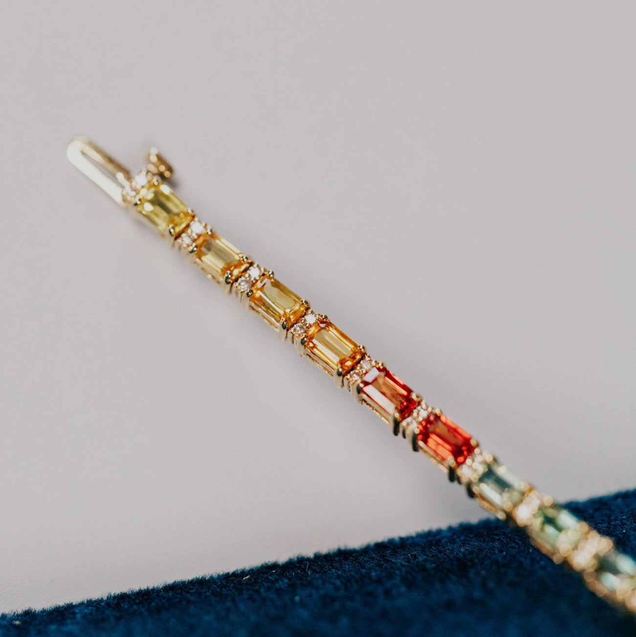 Close-up detail of the Rainbow Sapphire with Diamond Accent Tennis Bracelet in 14k yellow gold, highlighting the yellow, orange, and red emerald cut natural sapphires with diamond accents near the clasp, resting against a teal velvet surface.