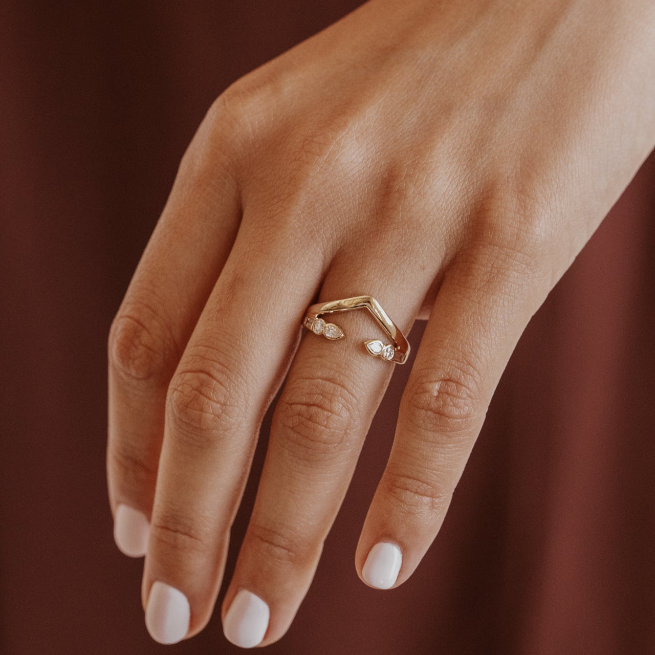 Close-up of a hand wearing a gold v wedding band with round and pear diamonds on a brown background