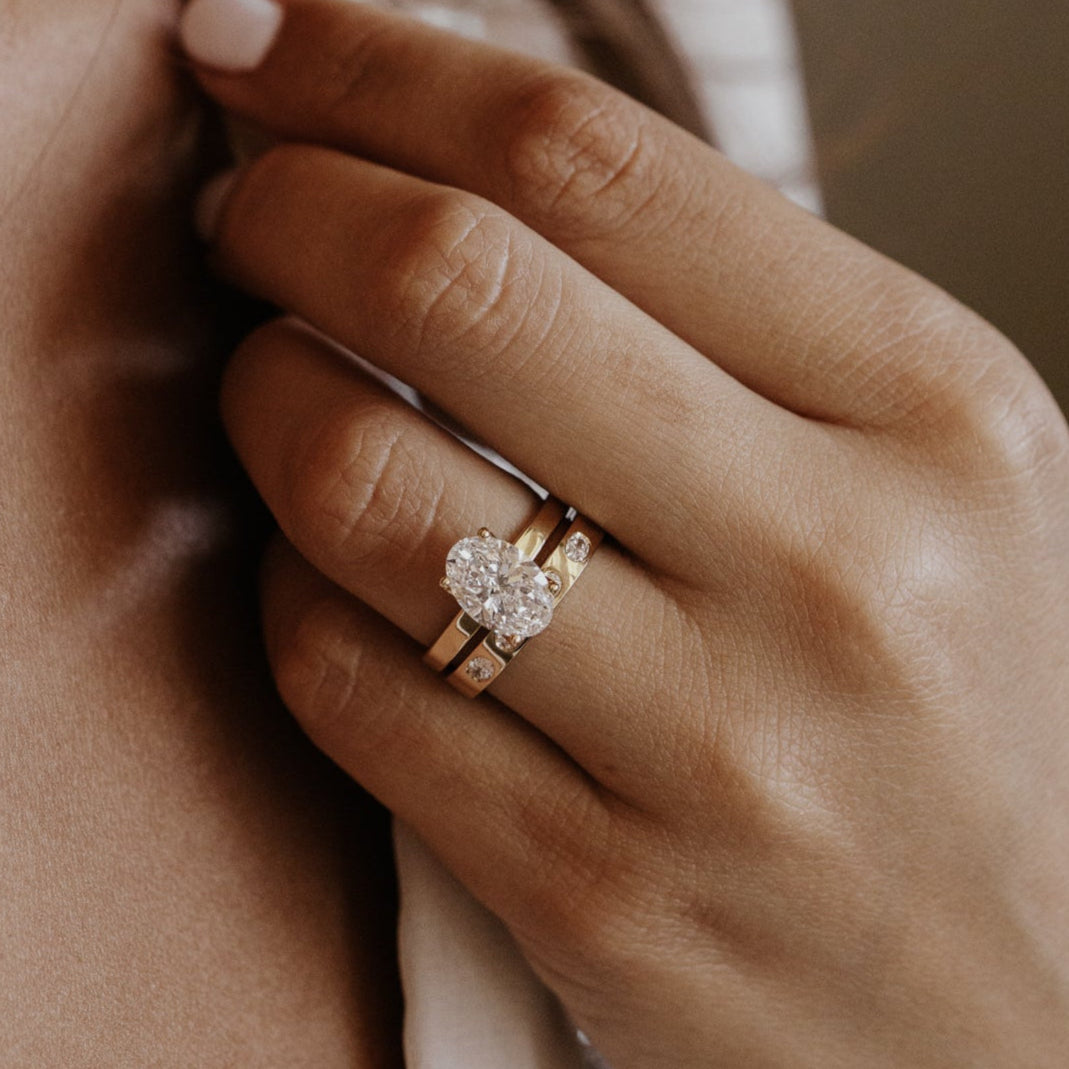 Close-up of a hand wearing a oval diamond ring with a gold oval diamond wedding band a blurred background