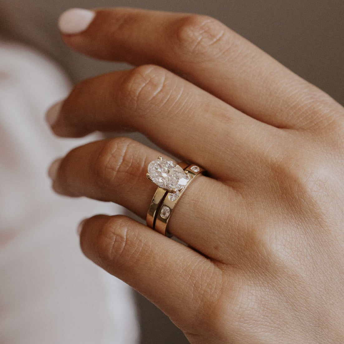 Close-up of a hand wearing a oval diamond engagement ring with a oval diamond gold band on a neutral background