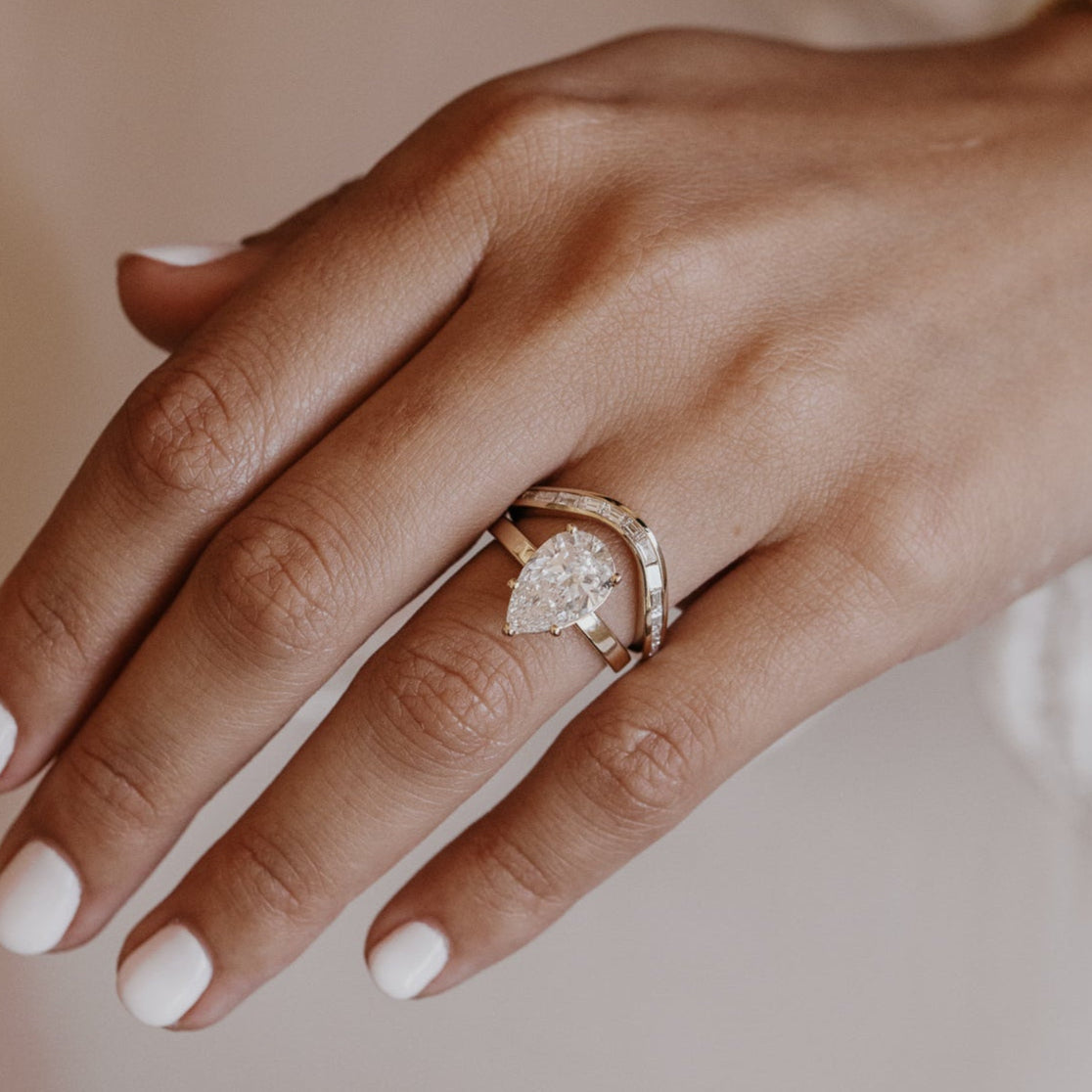 Hand wearing a pear engagement ring with a gold diamond wedding band on a blurred background