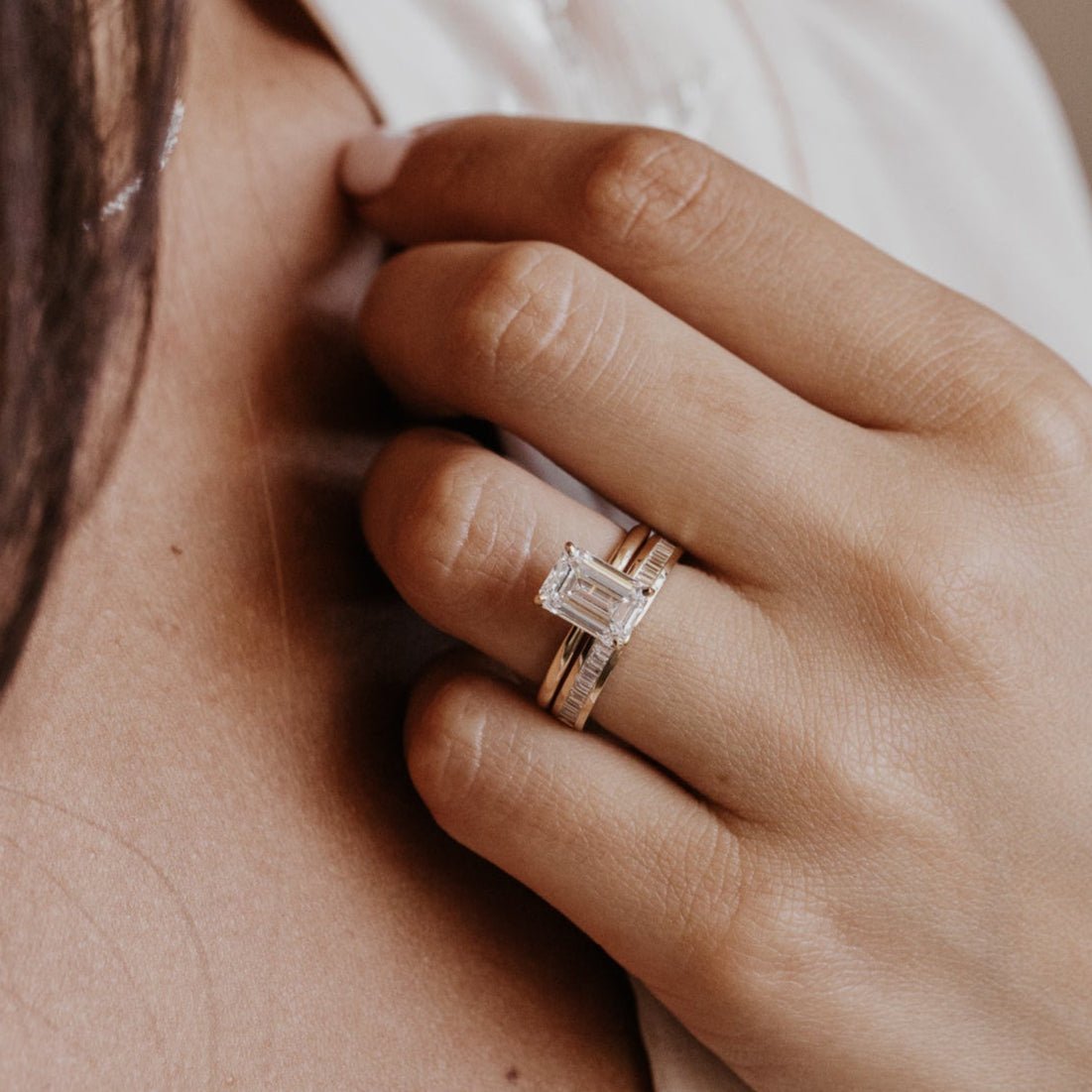 Close-up of a hand wearing a gold emerald cut diamond engagement ring with a diamond wedding band on a neutral background