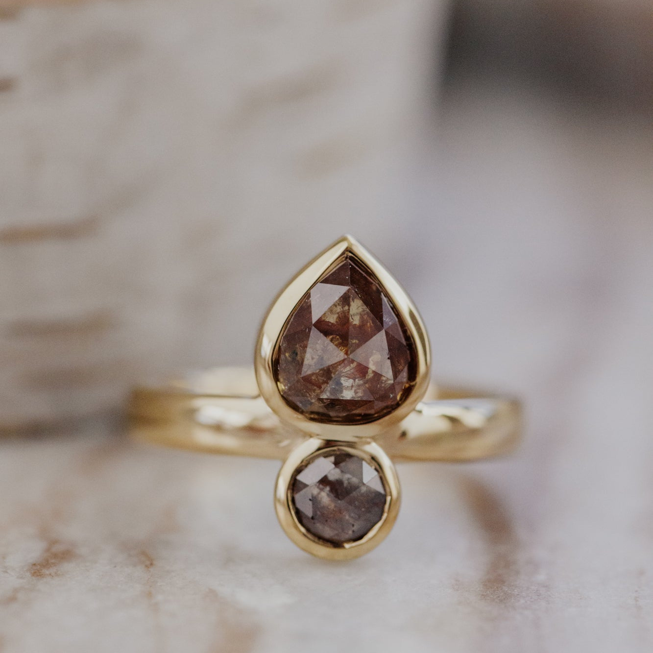 Gold Reese ring with a pear-shaped and round rustic diamond on a neutral background.