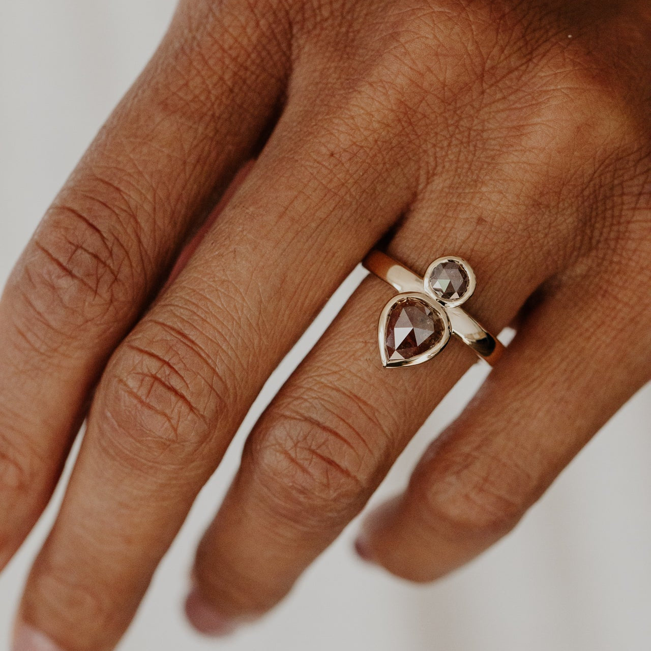 Hand wearing a Reese ring with pear and round rustic diamonds on a neutral background.