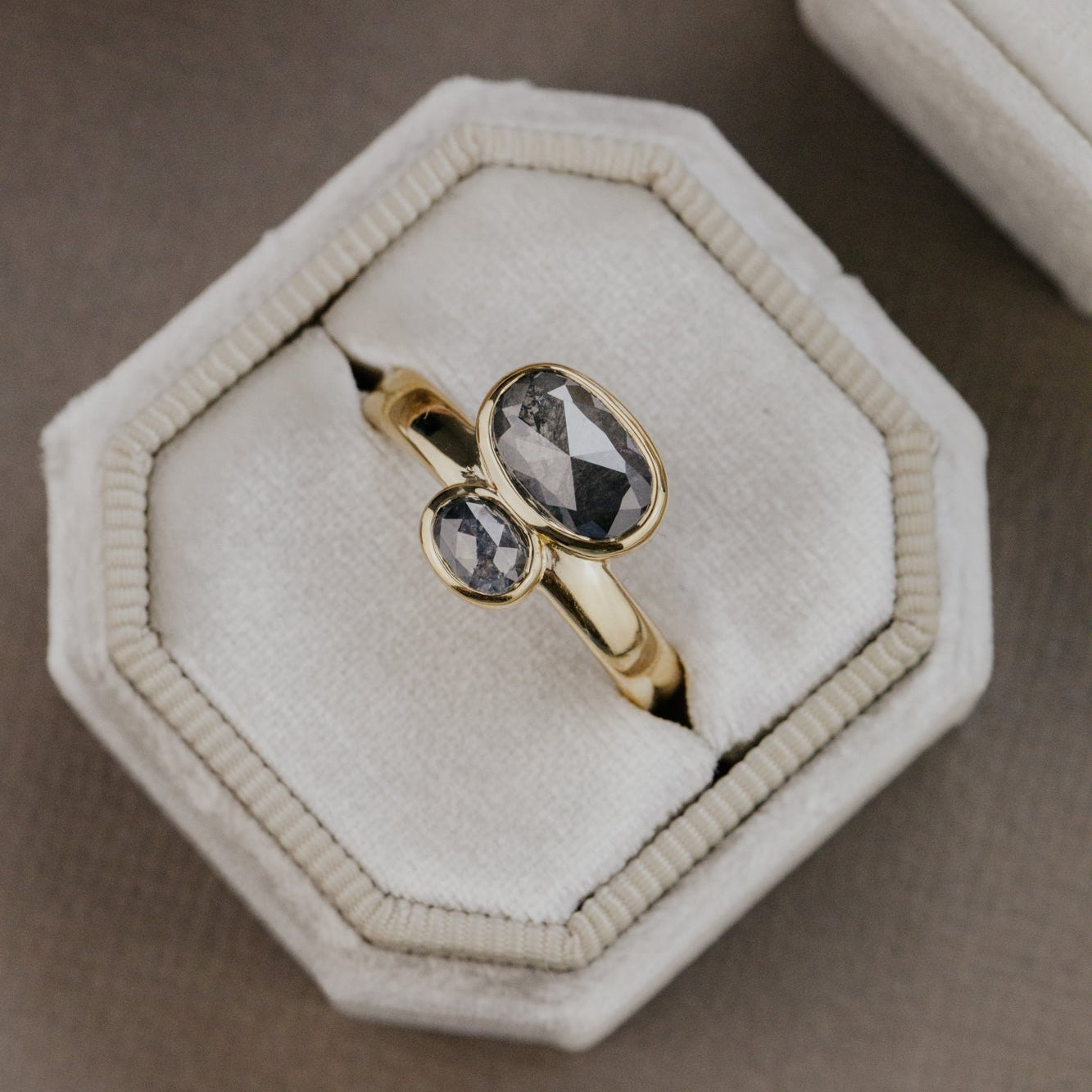 Gold ring with two gray rustic diamond gemstones on a beige hexagonal jewelry box.