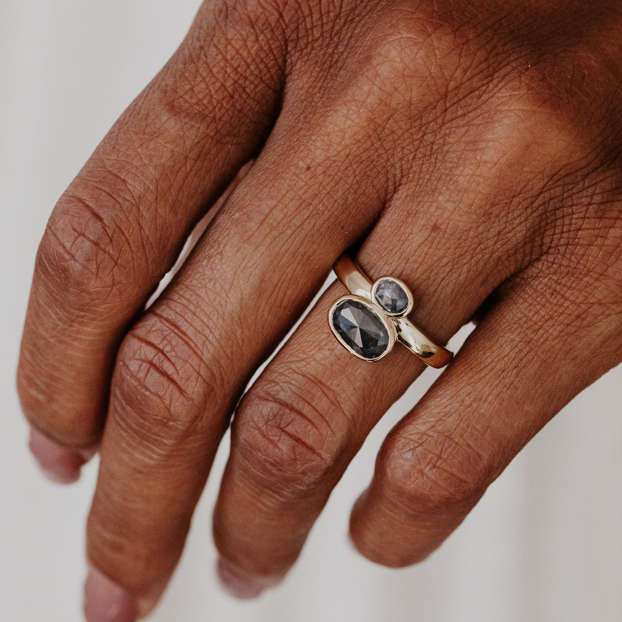 Hand wearing a gold ring with two dark rustic diamond gemstones on a neutral background.