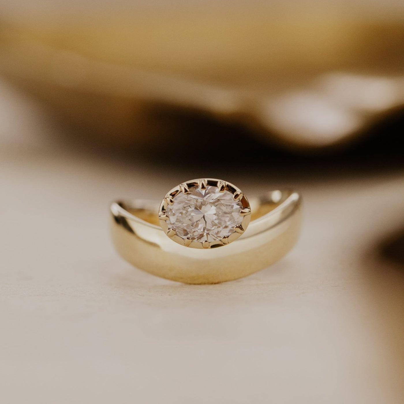 The Reflection gold pinky ring with an oval lab grown diamond on a blurred background.