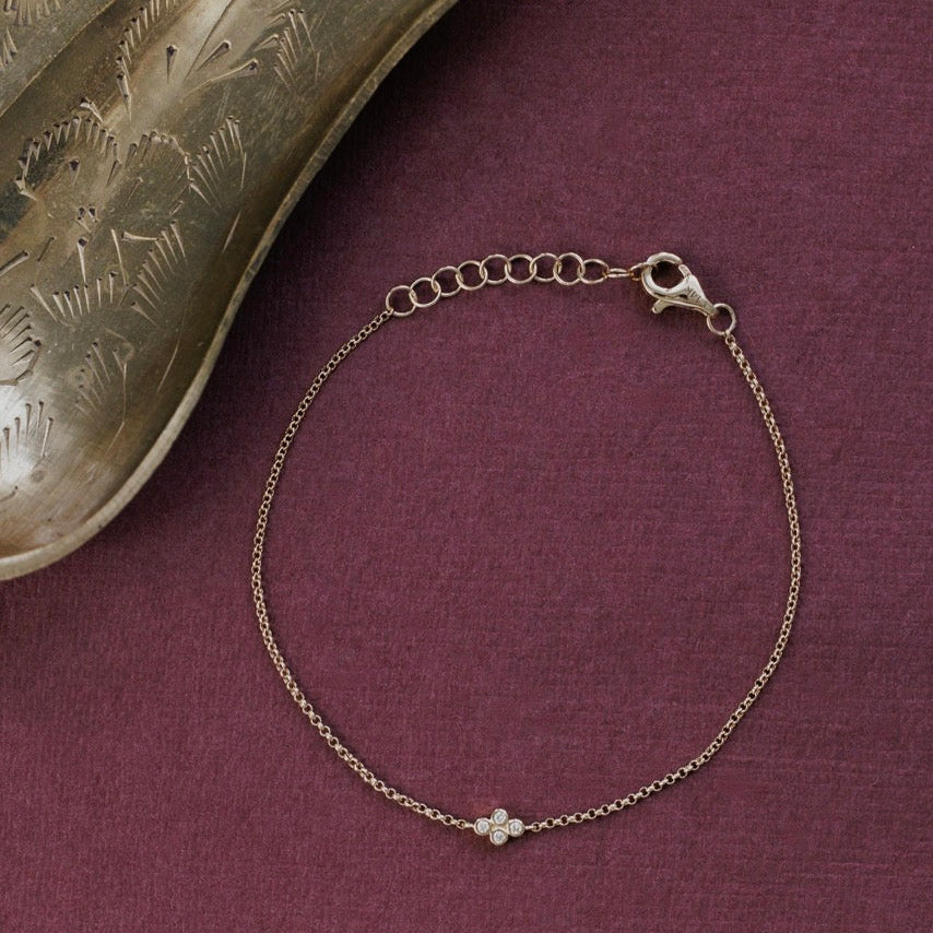 A delicate yellow gold chain bracelet with a diamond cluser, displayed on a textured dark purple surface next to a decorative metallic object.