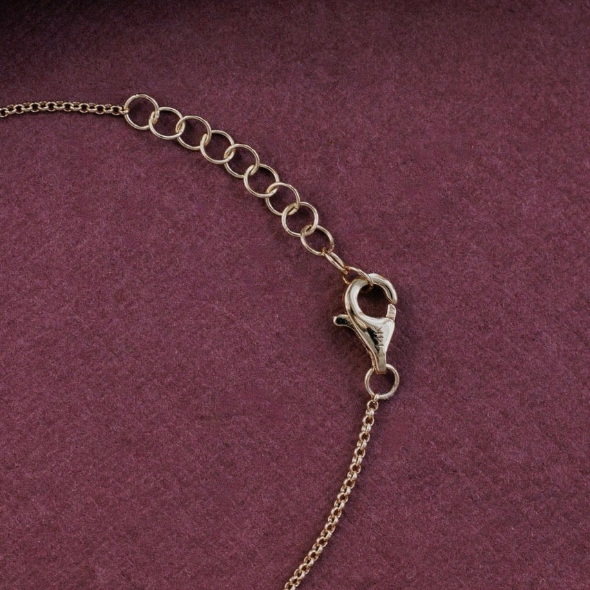 A close-up of a  yellow gold chain bracelet or necklace extender with a lobster clasp, laid on a dark purple textured surface with part of a metallic engraved object visible at the top.