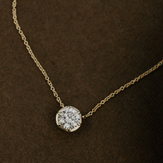 Gold necklace with a round lab grown diamond pendant on a brown background.