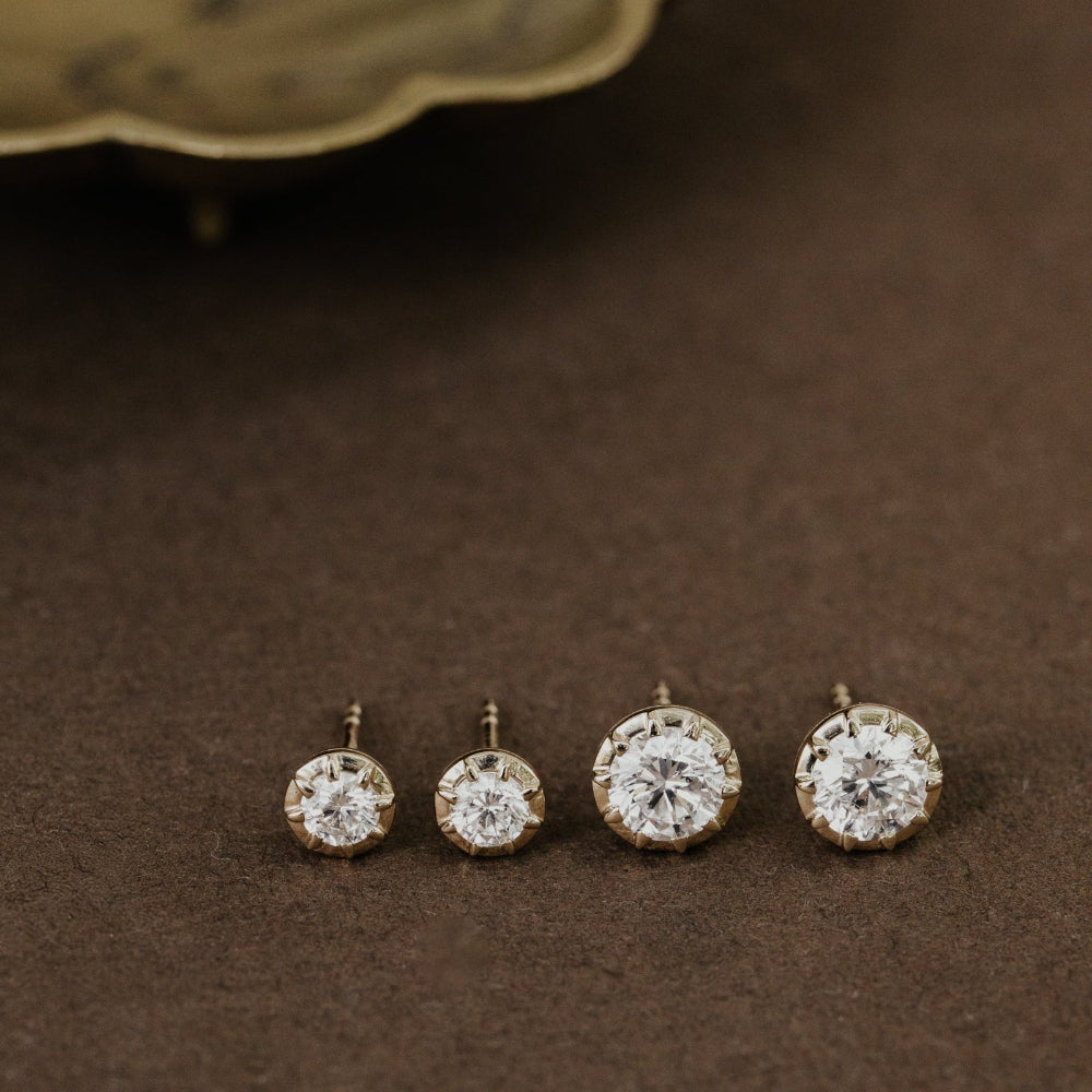Four lab grown diamond stud earrings on a brown surface with a decorative object in the background.